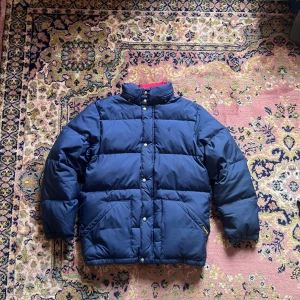 Mörkblå pufferjacka från Ralph Lauren - Snygg mörkblå pufferjacka från Ralph Lauren med broderad logga på bröstet och klassisk etikett vid fickan. Jackan har hög krage, tryckknappar och två stora fickor framtill. Perfekt för kalla dagar och riktigt stilren look.