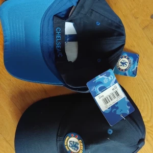 2nst Chelsea FC marinblå keps original 400 kr för båda 2 - Snygg marinblå keps från Chelsea FC med klubbens klassiska lejon-logga framtill. Kepsen har böjd skärm och justerbar passform. Tillverkad i bomull, perfekt för dig som vill visa ditt stöd för laget på matchen eller i vardagen.