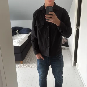 Svart overshirt från Uniqlo  - Svart overshirt från Uniqlo i enkel och clean design. Jackan har klassisk krage, knäppning framtill med svarta knappar och två bröstfickor med lock. Långärmad och i ett mjukt bomullstyg som passar perfekt till lager-på-lager. Storlek XS men jag har S och sitter nästan perfekt på mig