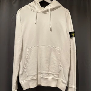 Vit hoodie från Stone Island - Vit hoodie från Stone Island med klassisk logga på ärmen. Tröjan har huva med snörning, magficka och ribbade muddar. Tillverkad i mjuk bomull, tyvärr har det blivit ett hål (bild 4), syns ej när man har på den och går säker att sy igen