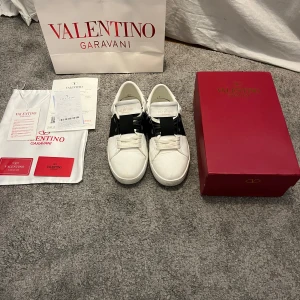 Valentino Garavani Open sneakers vit/svart - Säljer ett par Valentino Garavani Open sneakers i vitt skinn med bred svart rem över mitten. Klassisk rund tå, platt sula och ikoniska nitar baktill. Kommer med originalkartong, dustbag och kvitto. Perfekta för dig som vill ha en clean men lyxig look. Priset kan diskuteras!