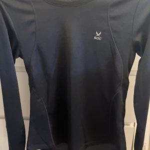 Svart långärmad träningströja SOC - Svart långärmad träningströja från SOC med diskret logga på bröstet. Tröjan har en slim passform och är tillverkad i ett mjukt och stretchigt syntetmaterial som andas, perfekt för träning eller löpning.