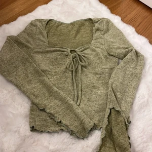 Grön långärmad topp med knyt från SHEIN - Superfin ljusgrön långärmad topp från SHEIN med söt knytning framtill och lätt croppad passform. Toppen har vågiga kanter vid ärmslut och nederkant, samt en mjuk och stretchig känsla tack vare viskos och elastan. Perfekt för dig som gillar trendiga detaljer!