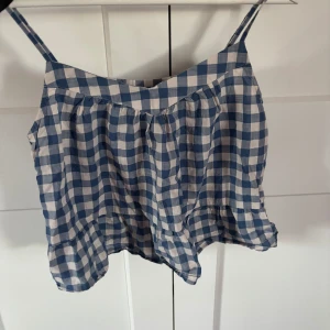 Blårutig linnetopp från GAP - Supersöt blårutig linnetopp från GAP med smala axelband och volangkant nedtill. Toppen är i tunn bomull och har en luftig passform, perfekt för varma dagar. Den klassiska rutiga designen i blått och vitt ger en fräsch och somrig vibe.