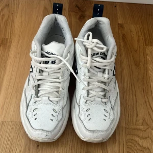 Vita New Balance sneakers med blå detaljer - Säljer ett par chunky vita New Balance sneakers med blå detaljer och klassisk N-logga på sidan. Skorna har snörning, rund tå och platt sula. Ovandelen är i syntet och skinn, med ventilerande hål och mörkblå hälflik. Perfekt för dig som gillar retrostil och sportig vibe.