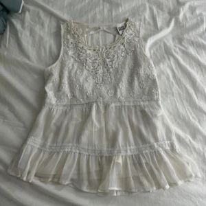 Vit spetsblus från Vero Moda - Supersöt vit blus från Vero Moda med broderade blommor och spetsdetaljer upptill. Blusen är ärmlös och har volang nertill samt transparenta partier som ger en romantisk vibe. Perfekt för sommarens alla tillfällen!