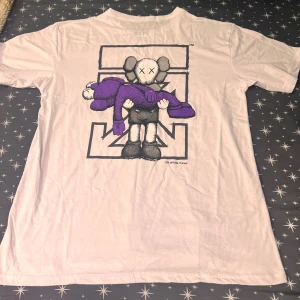 Off-White x Kaws vit t-shirt L - Vit t-shirt från Off-White x Kaws i storlek L, tillverkad i 100% bomull. Stor grafisk print på ryggen med Kaws-figur i svart, grått och lila. Framsidan har Off-White x André-logga tryckt över bröstet. Klassisk passform och rund hals.