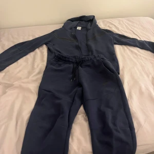 Mörkblå hoodie med dragkedja - Mörkblå hoodie från Nike med dragkedja framtill och huva. Tröjan har långa ärmar och en sportig look, perfekt för chill dagar eller när du vill vara bekväm. Materialet är mjukt och skönt, troligtvis bomull och polyester. Klassisk Nike-stil med diskret logga. Storlek på tröjan är Xl 158-170 cm / byxorna är Xs. Det är små knottrar på rumpan och lite på tröjan annars är den perfekt.