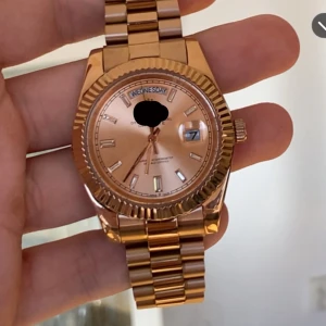 Day date 40 - Säljer en snygg guldig klocka med klassiskt länkarmband och rund urtavla. Klockan har dag- och datumvisning samt markerade index. Urtavlan och boetten är i matchande guldton, vilket ger en lyxig känsla.