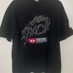Svart Diesel t-shirt med draktryck - Svart t-shirt från Diesel med coolt draktryck i grått på framsidan och stor Diesel-logga i vitt och rött både fram och bak. Klassisk rund hals och korta ärmar. Materialet är mjuk bomull, perfekt för en avslappnad streetstil.
