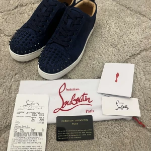 Christian Louboutin sneakers med nitar - Mörkblå sneakers från Christian Louboutin med klassisk röd sula och coola nitar på tån. Skorna har snörning, rund tå och är tillverkade i mocka med läderdetaljer invändigt. Perfekta för dig som vill sticka ut med exklusiv stil.
