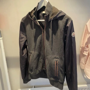 Moncler Jacka - Snygg grå hoodiejacka från Moncler med svarta nylonärmar, huva och dragkedja framtill. Jackan har två fickor med dragkedja och klassisk Moncler-logga på ärmen. 