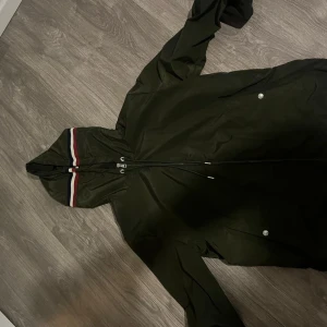 Grön vindjacka från Moncler med huva - Snygg mörkgrön vindjacka från Moncler med huva och dragkedja. Jackan har vita och röda ränder på huvan, två fickor med tryckknappar och Moncler-logga på ärmen. Perfekt för dig som vill ha en stilren och sportig look.