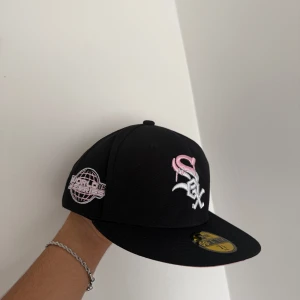 Svart New Era 59FIFTY White Sox keps - Snygg svart keps från New Era, modell 59FIFTY, med White Sox-logga i vitt och rosa broderi framtill. Sidopatch med 'World Series' i rosa och vitt. Platt skärm med rosa undersida. Tillverkad i 100% polyester, perfekt för dig som gillar sportig stil.