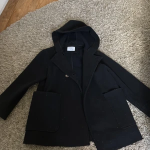 Svart skepparkavaj med huva från Zara - Svart skepparkavaj från Zara med huva och stora fickor framtill. Jackan har dubbelknäppning med fyra knappar och är tillverkad i ett ulligt material. Perfekt för dig som vill ha en klassisk look med modern twist.