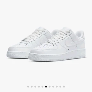 Nike Air Force 1 helvita sneakers - Klassiska Nike Air Force 1 sneakers i helvitt skinn med låg profil och rund tå. Skorna har perforerad tåbox, snörning och en tjock, platt sula. Perfekta för dig som gillar en clean och tidlös look.