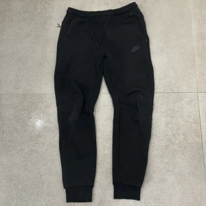 Svarta Nike mjukisbyxor med snörning - Svarta mjukisbyxor från Nike med snörning i midjan och ribbade muddar vid bensluten. Byxorna har en diskret Nike-logga på låret och dragkedjeficka bak. Perfekta för chill eller träning.