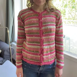 Randig kofta från Beyond retro - Sönder på baksidan! Säljer en färgglad stickad kofta från Beyond retro i rosa, beige, vitt och orange. Koftan har långa ärmar, knappar framtill och ett randigt mönster med små dekorativa pärlor. Perfekt för dig som gillar unika detaljer och mjuka material.