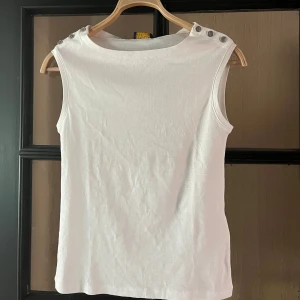 Vit ärmlös topp från Polo Ralph Lauren - Snygg vit ärmlös topp från Polo Ralph Lauren med rund halsringning. Tillverkad i mjuk bomull, perfekt för en clean och stilren look. Passformen är normal och toppen är enkel att matcha till olika outfits. Aldrig använd.