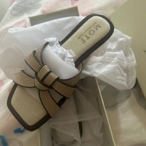 Beige sandaler från Mote med flätad detalj - Snygga sandaler från Mote i beige med flätad rem och fyrkantig tå. Skorna har platt sula och är tillverkade i textil och syntetmaterial. Perfekta för varma dagar och ger en stilren touch till din outfit.