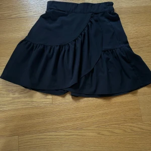 Svart omlottkjol med volang från Lindex - Svart kortkjol från Lindex i omlottmodell med bred volangkant. Kjolen har en elastisk midja som gör den bekväm och lätt att bära. Pris går att diskuteras!