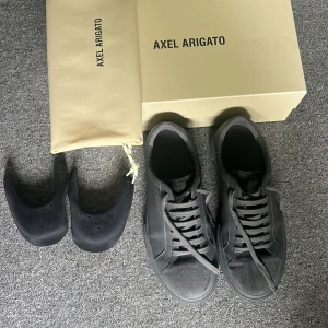 Svarta sneakers från Axel Arigato - Snygga svarta sneakers från Axel Arigato med stilren design. Skorna har ovandel i skinn och mocka, rund tå och platt sula. Klassisk snörning och diskret logga i guld på sidan. Perfekta för dig som gillar minimalistisk och modern stil.