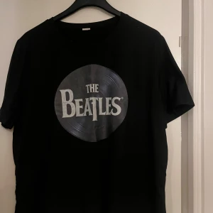 Svart The Beatles t-shirt - Svart t-shirt med stort The Beatles-tryck framtill i form av en vinylskiva. Klassisk rund hals och korta ärmar. Perfekt för dig som älskar musik och vill ha en ikonisk bandtröja i garderoben.