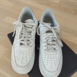 Nike Air Force 1 vita sneakers - Klassiska Nike Air Force 1 sneakers i vitt skinn med perforerad tå och snörning. Låg modell med vit sula och svart Nike-logga på sidan och hälen. Skickas i original skokartong (se foto).