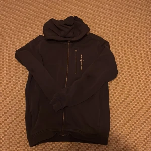 Svart hoodie från Sail Racing - Svart hoodie från Sail Racing med dragkedja framtill och broderad logga på bröstet. Tröjan har huva, lång ärm och en bröstficka med dragkedja. Mjuk insida och klassisk passform, perfekt för kyliga dagar samt bra inför vintern.