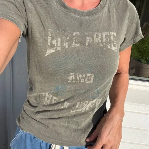 Grå/grön t-shirt från Juicy Couture - Cool grå/grön t-shirt från Juicy Couture med tryck i sliten vintage-stil och texten 'Live Free and...' framtill. Klassisk rund halsringning och korta ärmar. Mjuk bomullskvalitet och avslappnad passform, perfekt till jeans eller shorts. Stl.S 