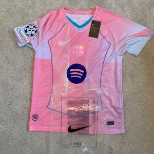 FC Barcelona rosa matchtröja Nike S - Snygg FC Barcelona fotbollströja från Nike i rosa och ljusblå toner. Tröjan har korta ärmar, V-ringning med blå kant och tryckta loggor från både Nike och Spotify. Tillverkad i lätt Dri-FIT-material som andas. Champions League-märke på ärmen.