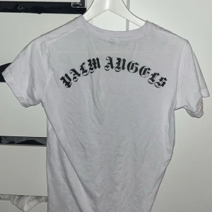 Vit Palm Angels t-shirt med tryck - Vit t-shirt från Palm Angels med svart tryck i gotisk stil över ryggen. Klassisk passform och rund halsringning, tillverkad i mjuk bomull. Perfekt för dig som gillar streetwear och vill sticka ut med en ikonisk logga.