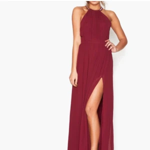 Vinröd långklänning med pärldetaljer EVE - Vinröd långklänning från EVE med halterneck och öppen rygg. Klänningen har pärl- och strassdetaljer längs banden i ryggen, hög slits framtill och är ärmlös. Tillverkad i ett lätt, flowy tyg som ger en elegant siluett.