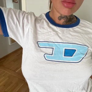 Retro Vit och blå t-shirt från Diesel - Vit t-shirt från Diesel med blåa detaljer och stor blå logga framtill. Klassiskt ”D” med blå ribbad kant vid hals och ärmslut. Skön passform och tillverkad i mjuk bomull.  Storlek L                              (Läs alltid min profil för villkor)