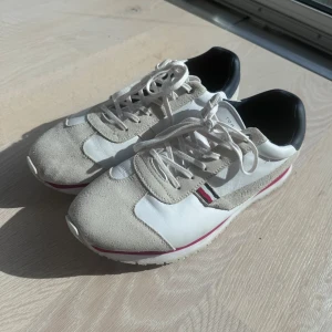 Tommy Hilfiger vita sneakers i skinn - Snygga vita sneakers från Tommy Hilfiger med detaljer i beige mocka och svart häl. Skorna har snörning, röd och marinblå rand på sidan samt en robust sula. Tillverkade i skinn och mocka för en clean och sportig look. Knappast använda
