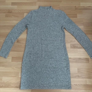 Grå klänning från Amisu - Mysig gråmelerad klänning från Amisu med lång ärm och hög krage. Modellen är rak och kort, perfekt för lager på lager. Materialet är mjukt och stretchigt, vilket gör den superbekväm. Enkel och stilren design som funkar till många olika looks. Använd en gång.