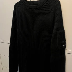Svart stickad tröja från C.P. Company - Svart ribbstickad tröja från C.P. Company med deras ikoniska linsdetalj på ärmen. Tröjan har rund halsringning och är i jätte bra skick då jag ändrat använt den fåtal gånger och därav priset.