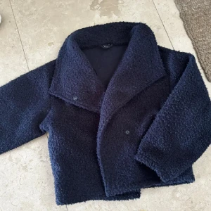 Mörkblå teddyjacka med bred krage - Säljer en mörkblå teddyjacka med bred krage och knäppning framtill. Jackan har en boxig passform och är tillverkad i mjukt, fluffigt material som ger en cool och avslappnad look. Perfekt för dig som gillar trendiga och bekväma ytterplagg.