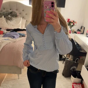 Ljusblå blus med prickar och volang från hm - Säljer en söt ljusblå blus med diskreta prickar, volangdetaljer längs axlarna och knappar framtill. Blusen har långa puffärmar med smock vid ärmslut och v-ringad hals. Från h&m