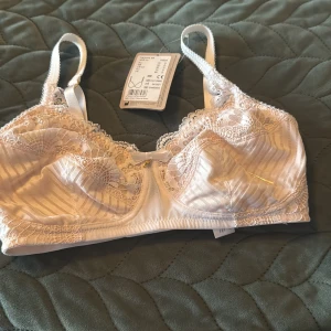 Vit bh från Amoena - Säljer en vit bh  från Amoena med fina spetsdetaljer och diskreta ränder. Bh har justerbara axelband, liten rosett framtill och mjuka kupor med spets. Perfekt för dig som gillar romantisk och feminin stil.