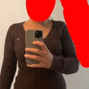 Brun kabelstickad tröja Hollister - Mysig brun kabelstickad tröja från Hollister med v-ringning och långa ärmar. Tröjan har ribbade muddar och en liten broderad logga på bröstet. Perfekt för lager-på-lager under hösten och vintern. 2000s style.