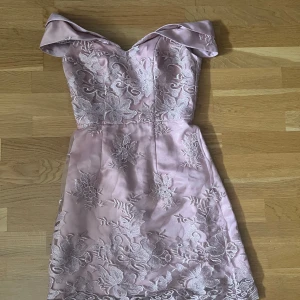 Ljusrosa spetsklänning från Chi Chi London - Supersöt ljusrosa kortklänning från Chi Chi London med offshoulder och v-ringad hals. Klänningen har vackra blommiga spetsdetaljer och är gjord i glansigt satinliknande material. En axelrem har lossnat men kan lätt åtgärdas.