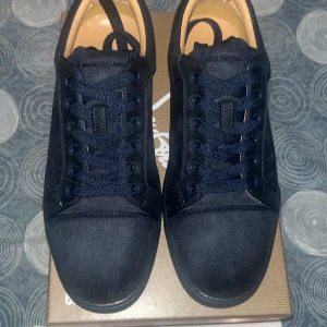 Christian Louboutin svarta sneakers mocka - Snygga svarta sneakers från Christian Louboutin i mocka med klassisk röd sula och diskret logga undertill. Skorna har rund tå, platt sula och snörning. Perfekta för dig som vill ha en lyxig och stilren look med en ikonisk touch.