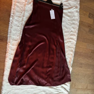 Vinröd satinkjol från Stockh lm - Snygg vinröd långkjol i satin från Stockh lm. Kjolen har en slät, glansig yta och en diskret slits framtill. Detalj med metallring i midjan och dragkedja i sidan. Perfekt för dig som gillar stilrena och eleganta plagg med en twist.