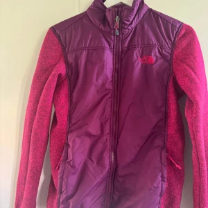 Vinröd vindjacka från The North Face - Säljer en vinröd vindjacka från The North Face med hög krage och hel dragkedja framtill. Jackan har en sportig look med paneler i olika nyanser av rött och diskret logga på bröstet. Perfekt för dig som gillar en aktiv stil och vill ha något lätt och smidigt.