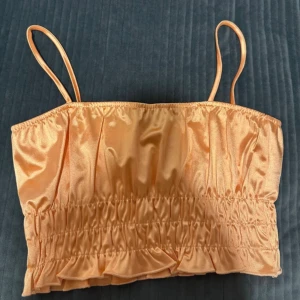 Persikofärgad satin linnetopp - Snygg persikofärgad linnetopp i glansigt satinmaterial med smala axelband och rynkade detaljer. Perfekt för varma dagar och somriga outfits. Croppad modell som ger en trendig look.