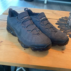 Nike Vapormax Flyknit svarta sneakers - Säljer ett par svarta Nike Vapormax Flyknit sneakers med stickad ovandel och synlig luftsula. Skorna har snörning och en sportig, modern look med diskret Swoosh på sidan. Perfekta för dig som gillar streetwear och vill ha riktigt sköna och lätta sneakers.