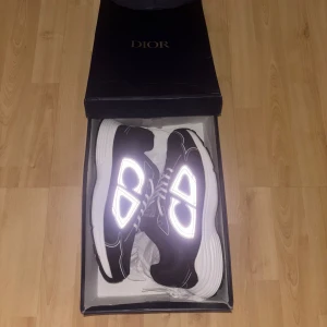 Svarta Dior sneakers med reflekterande detaljer - Säljer ett par svarta Dior sneakers med vita snören och fet reflekterande CD-logga på sidorna. Skorna har en chunky vit sula. Grymma detaljer och riktigt snygg siluett, perfekta för dig som vill sticka ut.