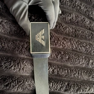 Svart skärp från Emporio Armani - Snyggt svart bälte från Emporio Armani med stilren rektangulär metallspänne och logotyp. Tillverkat i äkta skinn och har en klassisk, minimalistisk look som passar till jeans eller kostym. Made in Italy och känns riktigt lyxigt.