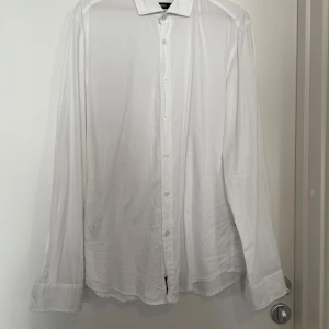 Vit skjorta från Hugo Boss XL - Klassisk vit skjorta från Hugo Boss i slim fit-modell. Skjortan har långa ärmar, knappar framtill och en stilren krage. Perfekt för dig som gillar en clean och tidlös look. Tillverkad i mjuk bomull för skön känsla hela dagen.